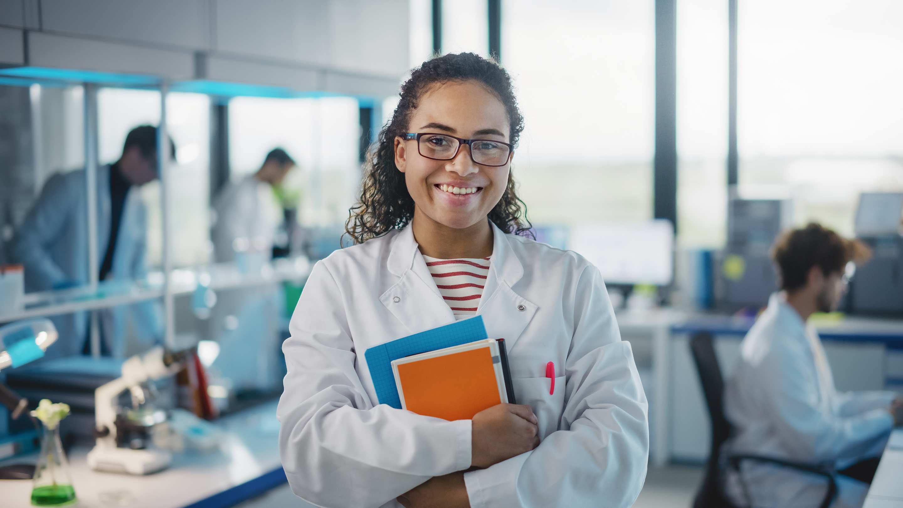 Medical Science Laboratory: young med student wearing white coat and glasses, holds text books, smiling, looking at camera