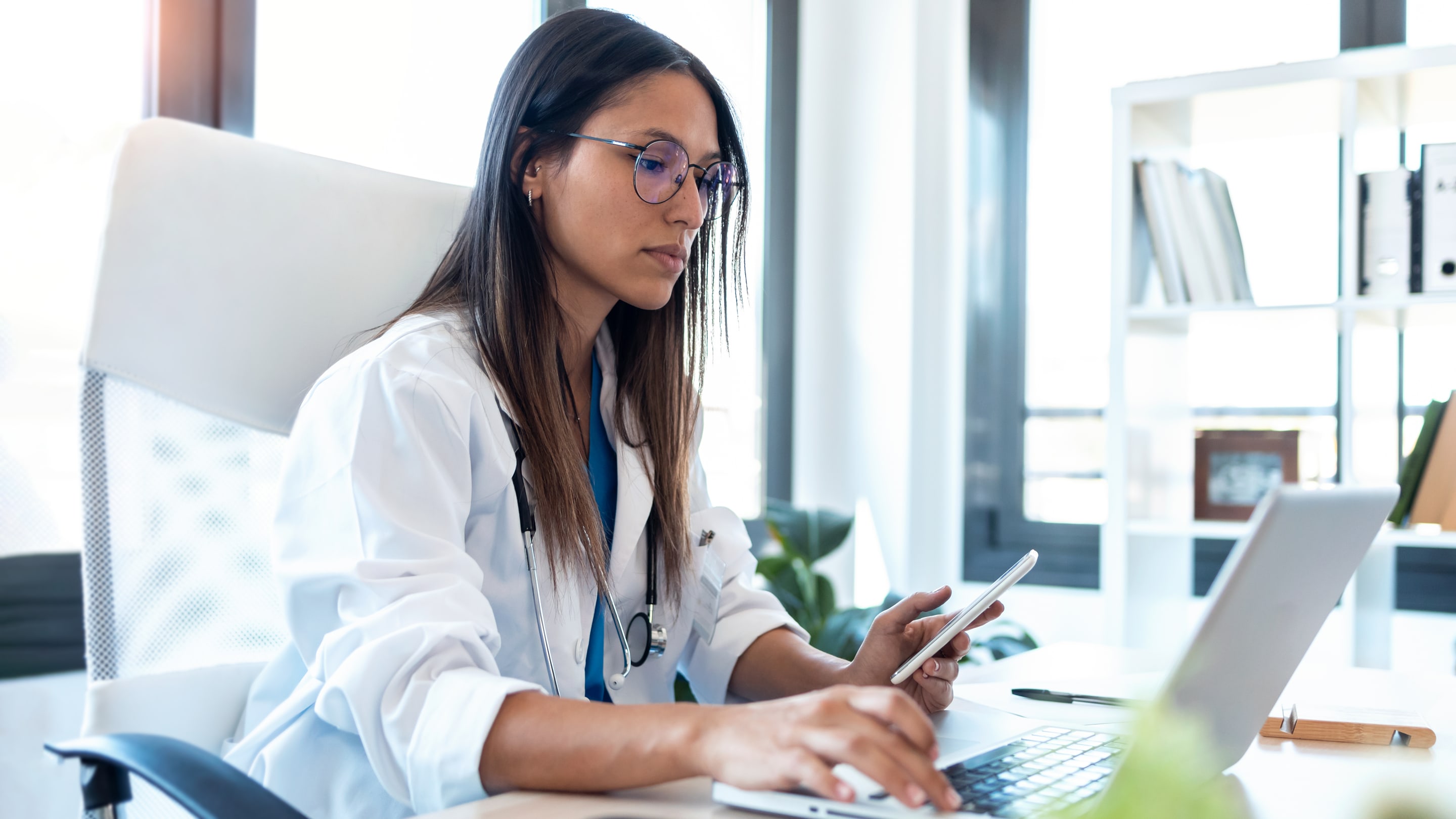 Image of confident young female doctor using her mobile phone while working on laptop in the consultation.