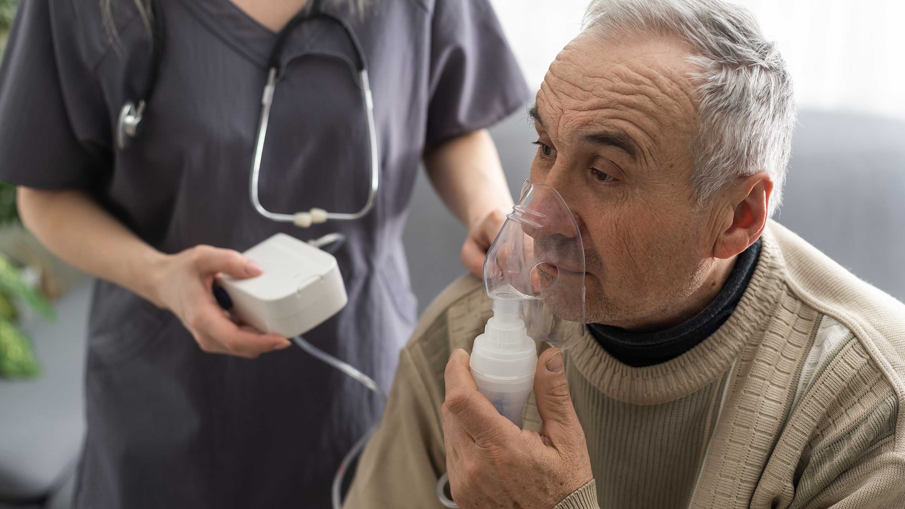 Senior adult male patient using asthma COPD breath nebulizer and mask as clinician stands nearby.