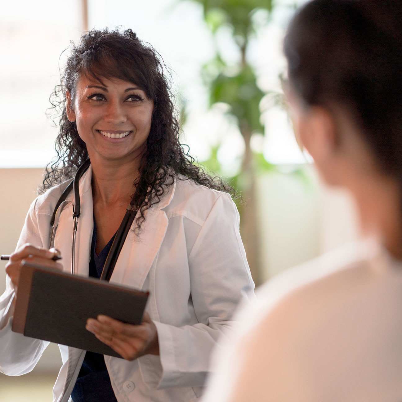 Young, female doctor in a white coat holds a clipboard and smiles at a patient.