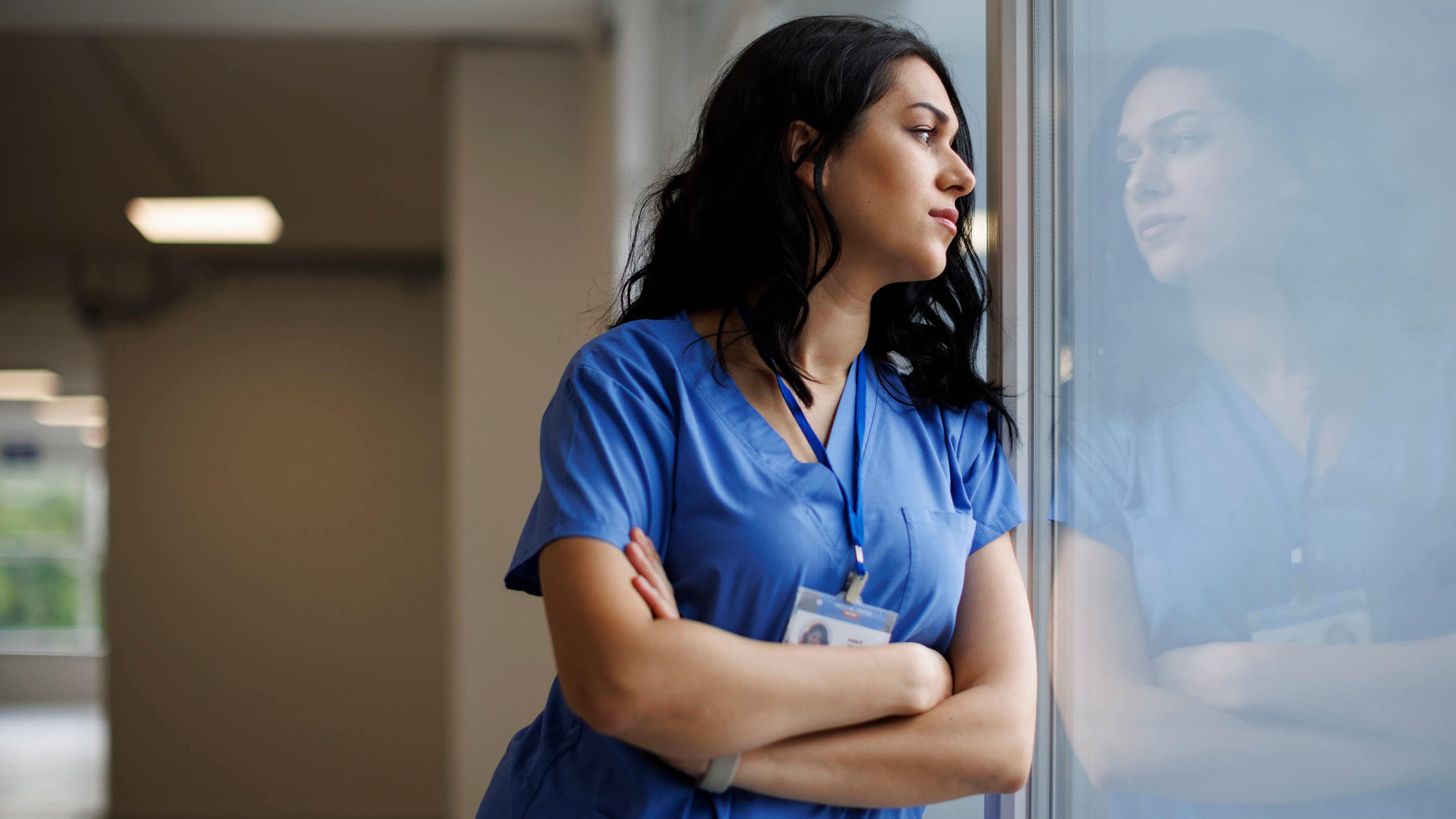 Female healthcare working looks out reflective window with her arms crossed and has a neutral face.