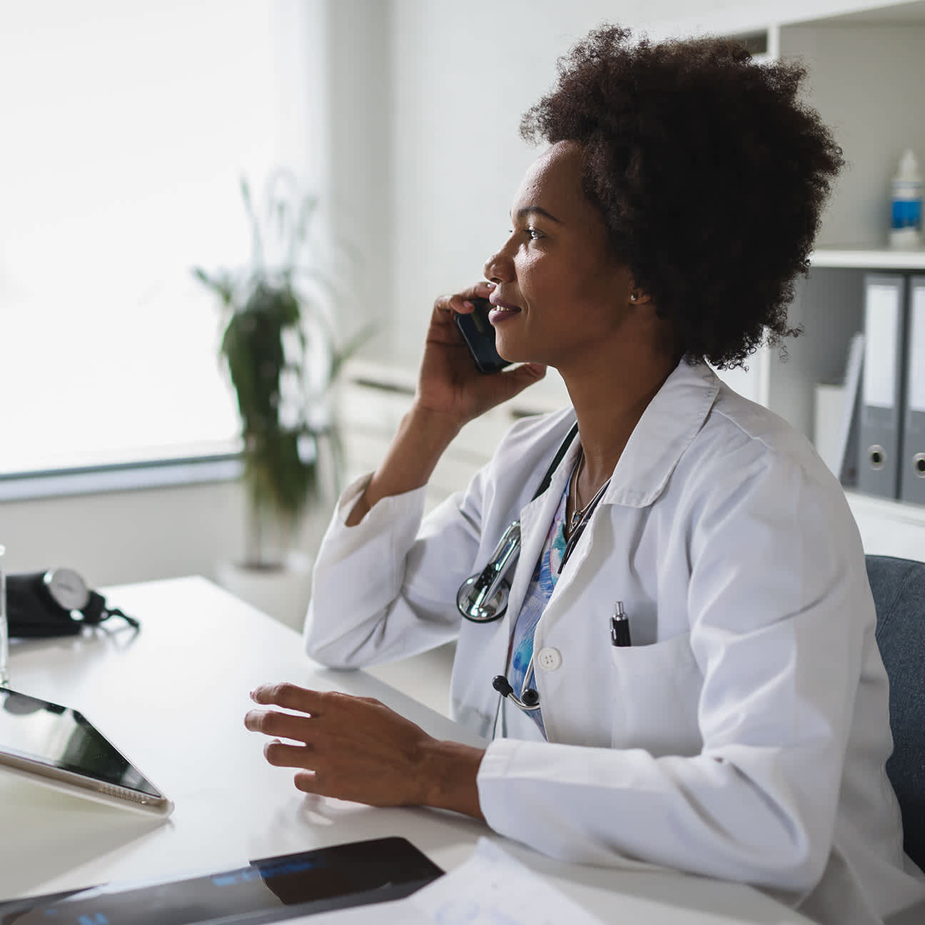A Black female doctor speaks on the phone at her desk.