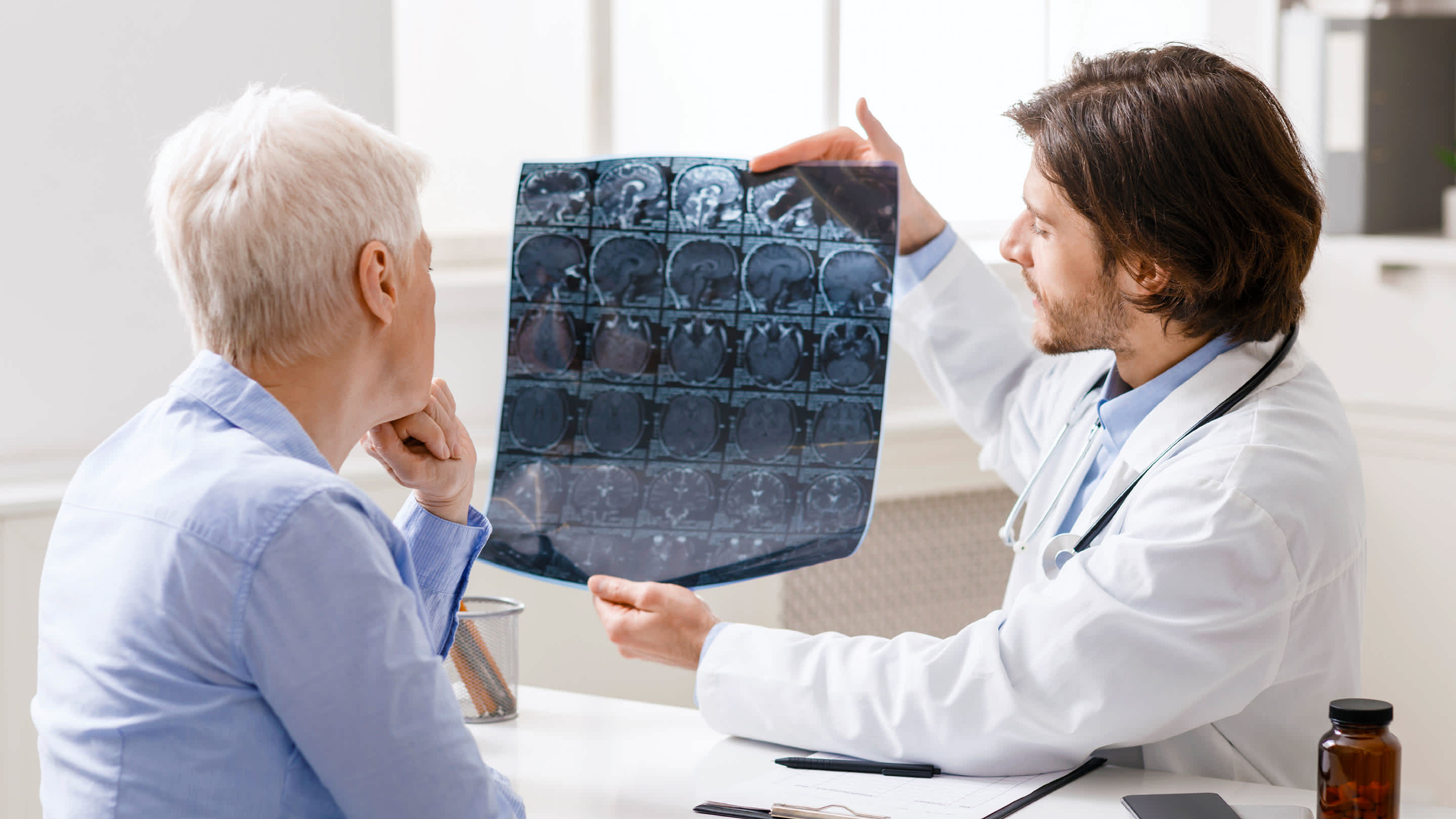 A young male doctor reviews brain imaging results with an older male patient in an office setting.